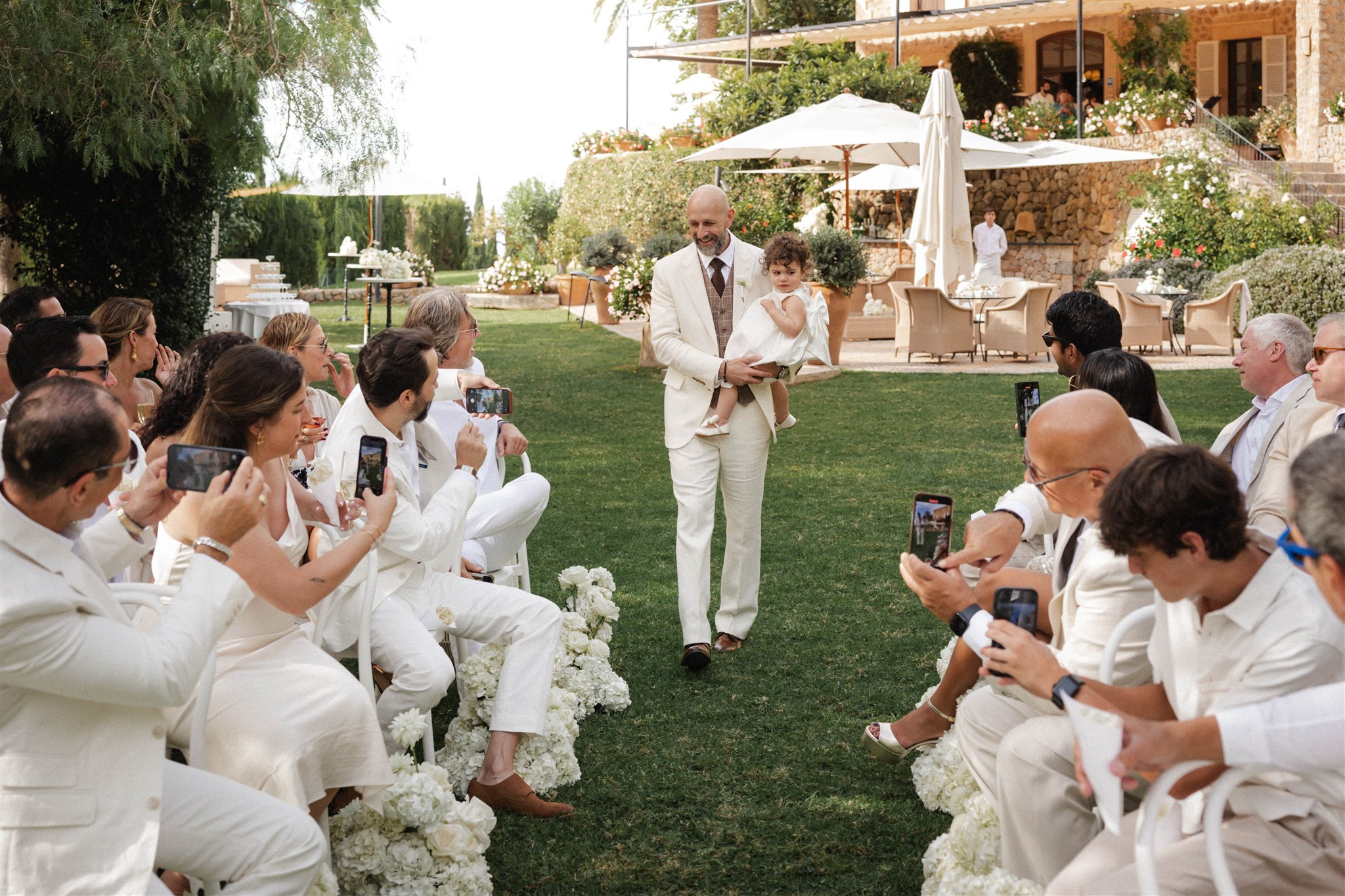 Groom holding toddler daughter while bride approaches at mountain wedding ceremony.
