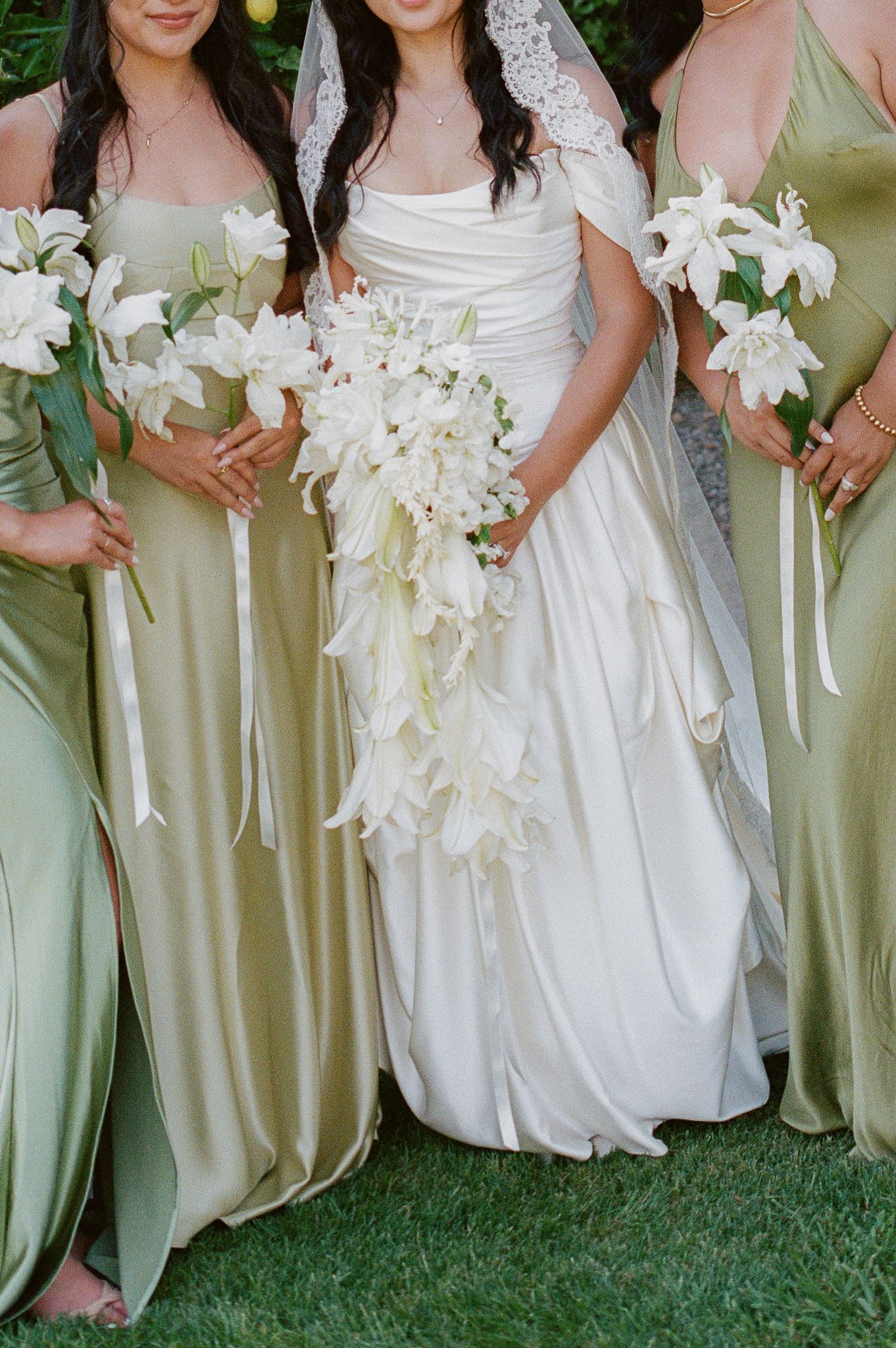 Bride with bridesmaids in sage green satin dresses holding white lily bouquets