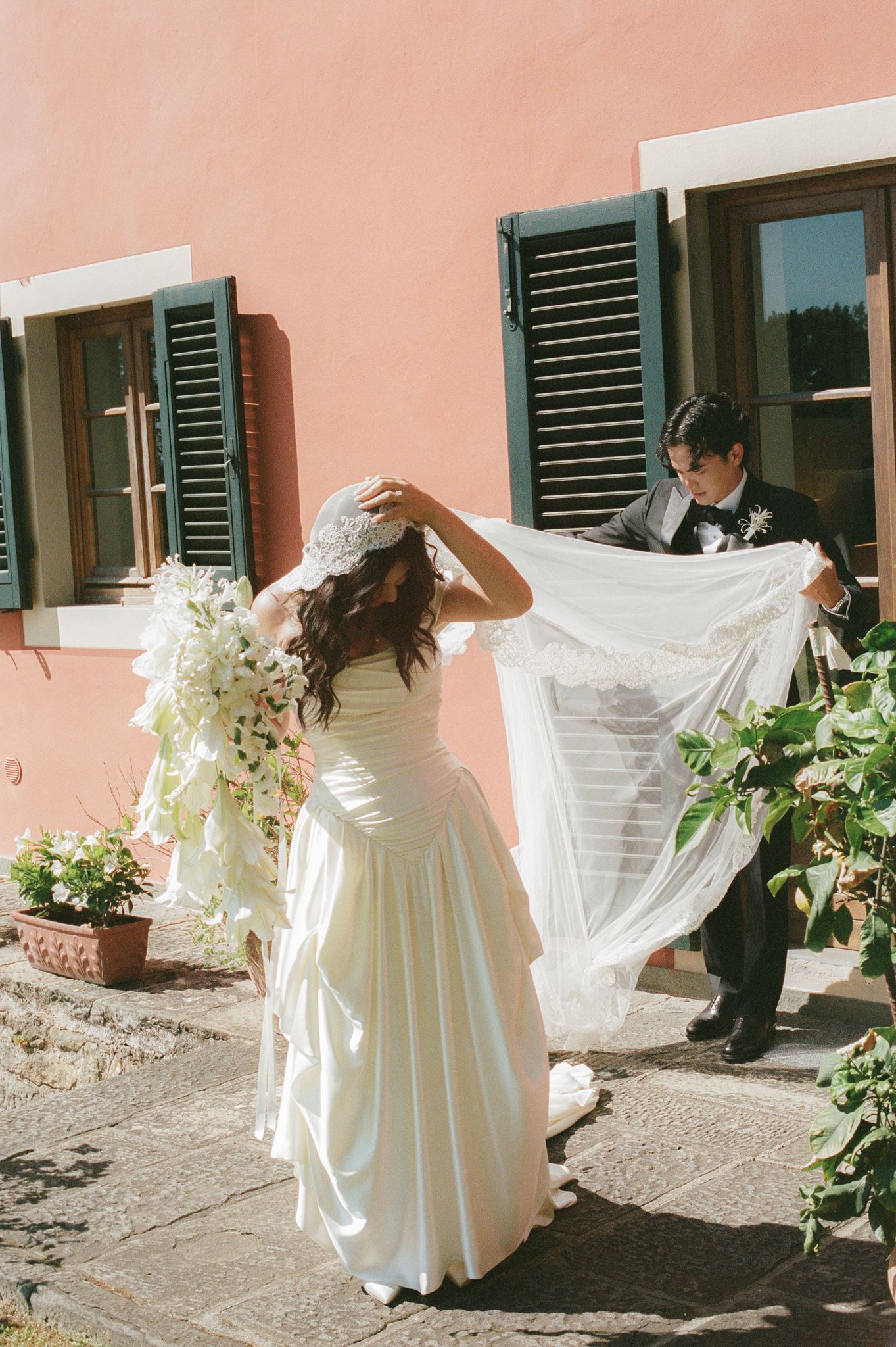 Bride in a ruched wedding gown and lace veil holding a cascading white bouquet as the groom lifts her veil at an Italian villa