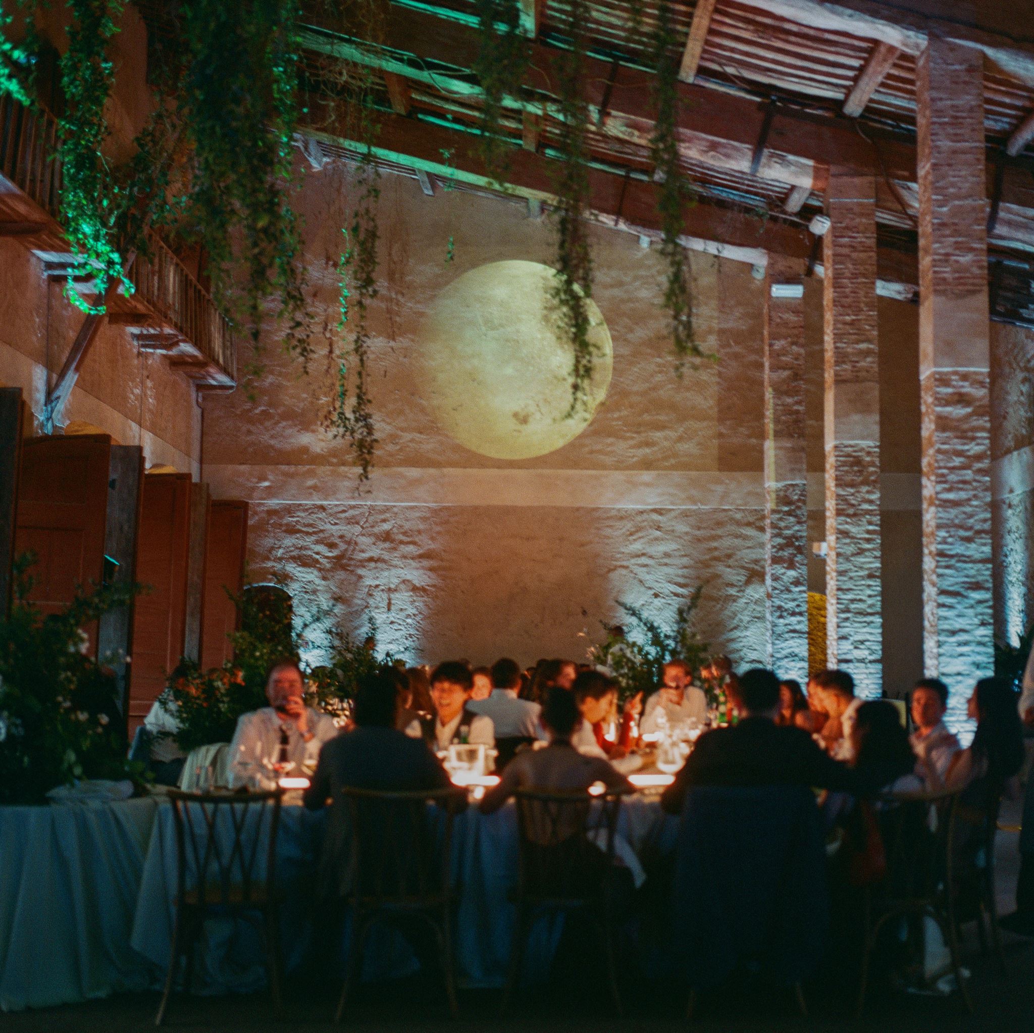 Modern wedding reception tables with white linens, sculptural greenery, linear lighting, and a moon projection backdrop