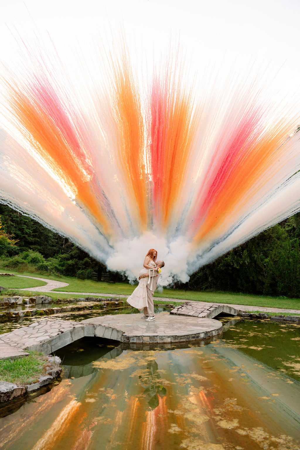 Bride and groom embrace beside a pond as orange, pink and white smoke bombs burst behind them at their Mount Olympus wedding in Greece.