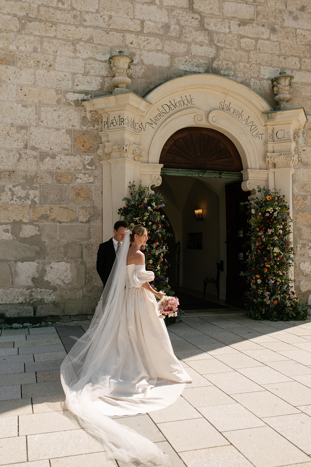 Bride Teresa in a flowing white wedding gown outside a historic stone church in Austria.