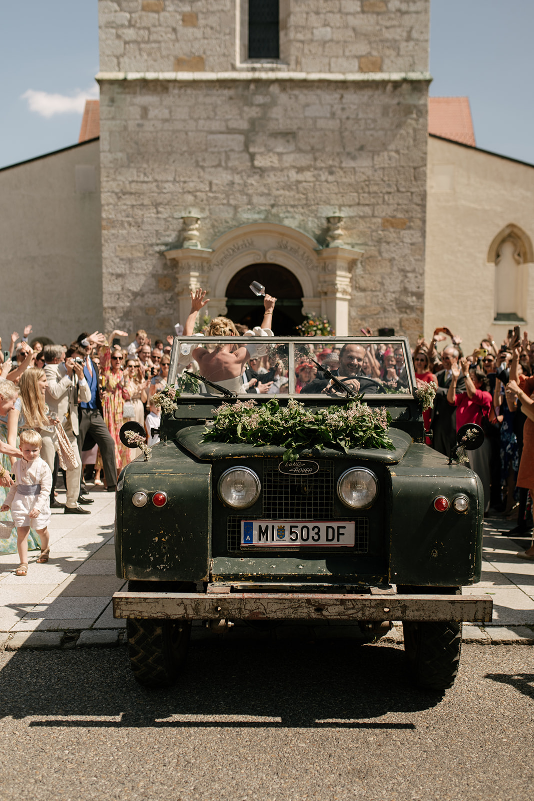 Teresa and Moritz celebrate their wedding with guests outside a historic Austrian church as a vintage car waits nearby.