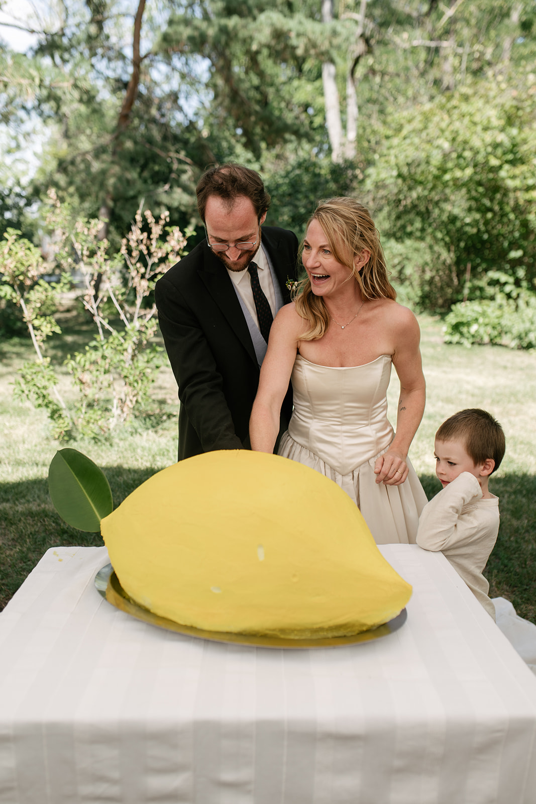 Bride and groom with their child behind a whimsical giant lemon-shaped wedding cake at their Austrian countryside reception.