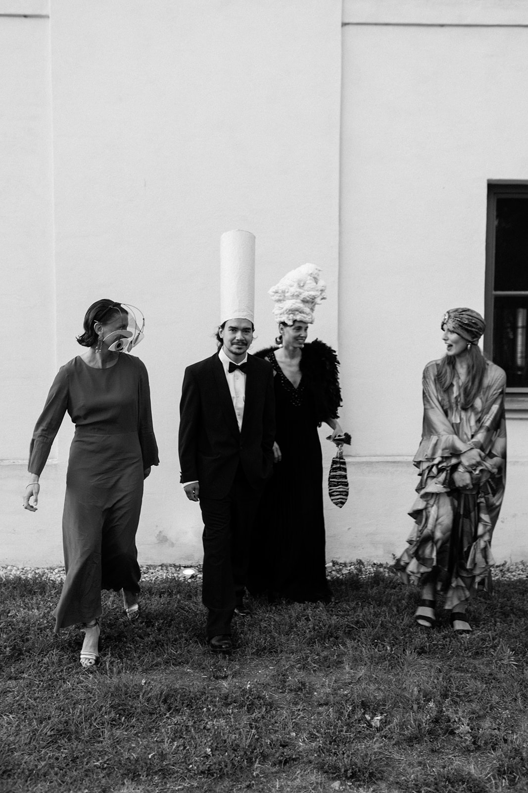 Black-and-white photograph of wedding guests in elegant eveningwear and elaborate sculptural headpieces.