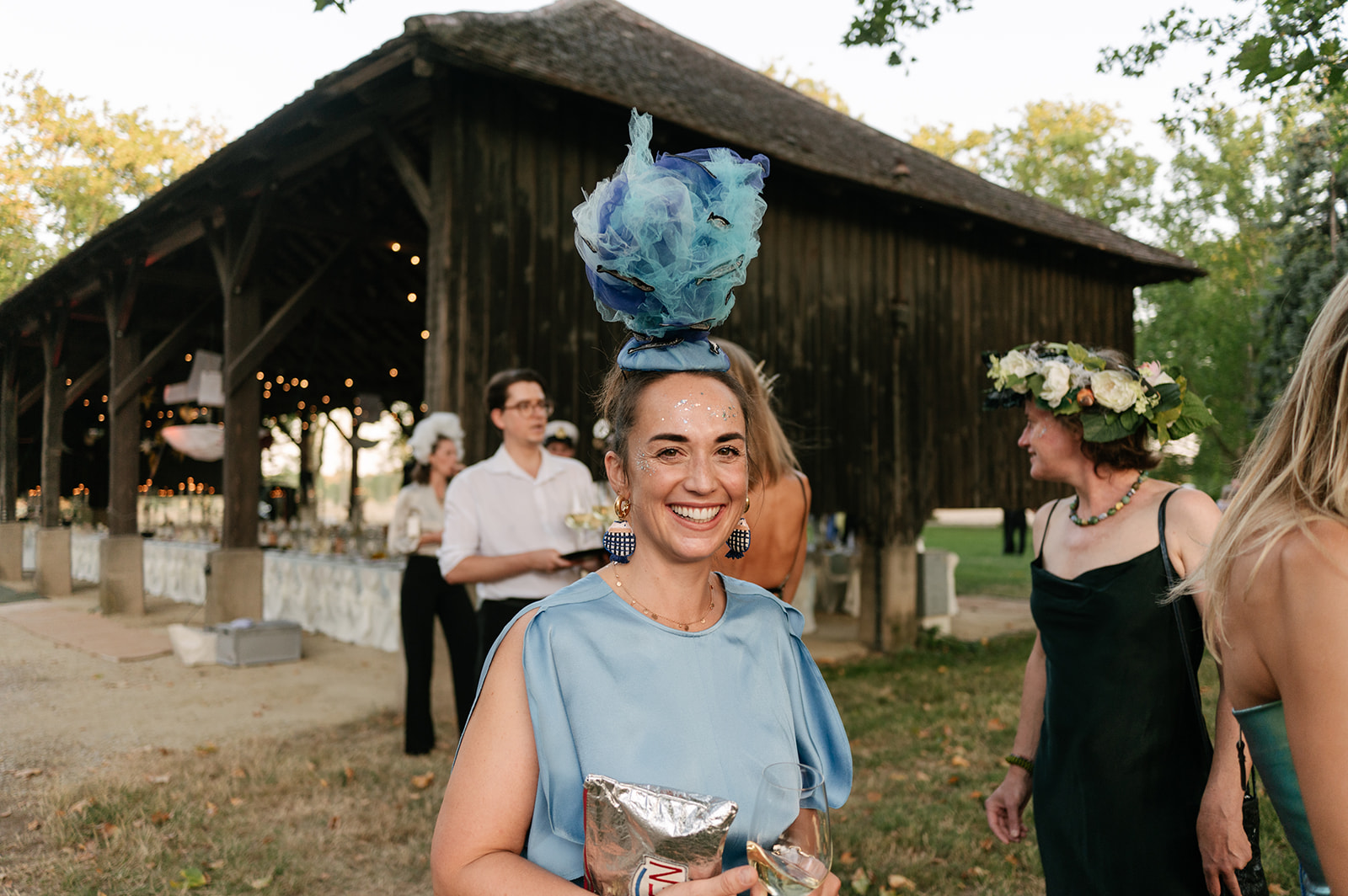 Smiling wedding guest wearing a bright blue dress and oversized sculptural blue headpiece at a rustic barn reception.
