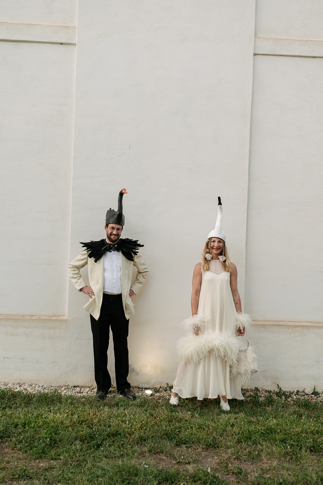 Bride and groom wearing whimsical handmade swan headpieces and artistic wedding attire against a minimalist white wall.