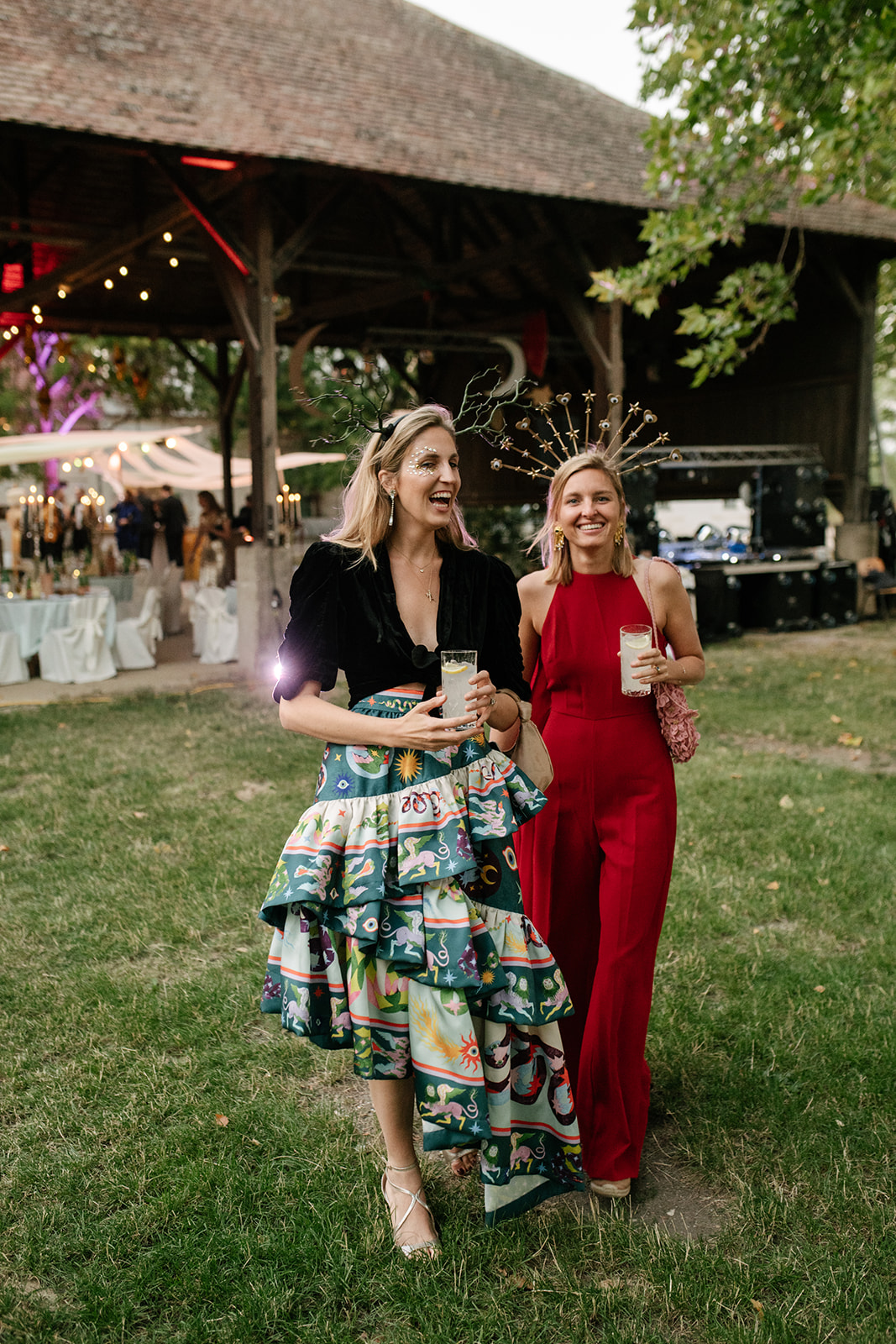 Two wedding guests in colorful floral and red dresses smiling together at an outdoor barn wedding reception.